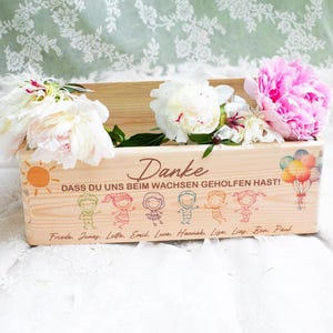 May include: A wooden planter box filled with white and pink peonies. The box has a sun and cartoon figures with the German text "Danke" and "Dass du uns beim wachsen geholfen hast!" with names below. A bouquet of balloons is also depicted.