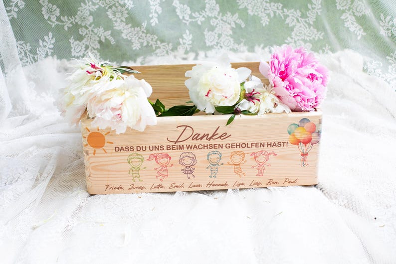 May include: A wooden planter box filled with white and pink peonies. The box has a sun and cartoon figures with the German text "Danke" and "Dass du uns beim wachsen geholfen hast!" with names below. A bouquet of balloons is also depicted.