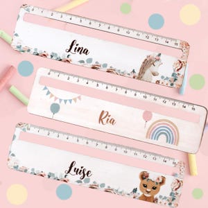 Könnte beinhalten: Drei Holzlineale mit Blumendesigns und personalisierten Namen: Lina, Ria und Luise. Jedes Lineal zeigt eine andere Illustration: ein Einhorn, einen Regenbogen mit einem Ballon und einen Löwen.