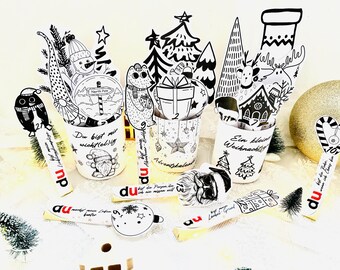 May include: A collection of black and white Christmas-themed decorations, including cups with illustrations of a snowman, bear, and house. The decorations also include small Christmas trees and ornaments. Some items have text in German.