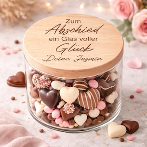 Könnte beinhalten: Ein Glasbehälter gefüllt mit herzförmigen Pralinen und Süßigkeiten, bedeckt mit einem Holzdeckel. Auf dem Deckel stehen die Worte "Zum Abschied ein Glas voller Glück Deine Jasmin". Die Pralinen sind in Braun-, Weiß- und Rosatönen gehalten.