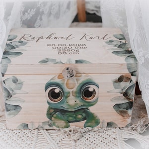 Peut inclure: Une boîte à souvenirs en bois avec une finition en bois clair et une illustration de tortue verte et brune. La boîte est décorée de feuilles vertes et porte le texte "Raphael Karl 23.08.2023 09:30 Uhr 3380g 53 cm" sur le couvercle.