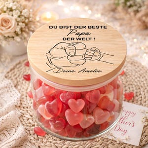 Könnte beinhalten: Ein Glas mit herzförmigen Süßigkeiten, bedeckt mit einem Holzdeckel. Der Deckel trägt den Text "DU BIST DER BESTE Papa DER WELT!" und eine Zeichnung von zwei Händen. Eine Karte mit "Happy Mother's Day" ist sichtbar.