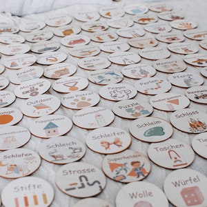 May include: A set of 50 round wooden tokens with various illustrations and German words. The tokens are arranged in a grid pattern on a light brown surface. Some of the words include "Kreide", "Matador", "Outdoor", "Puppenhaus", "Schlafen", "Schleich", "Schleifen", "Schleim", "Schloss", "Schminke", "Stifte", "Strasse", "Superhelden", "Taschen", "Tierarzt", "Tiere", "Wolle", "W&uuml;rfel", and "Zahlen".