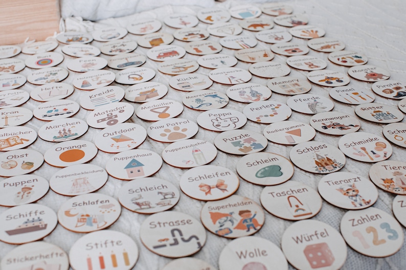 May include: A set of 50 round wooden tokens with various illustrations and German words. The tokens are arranged in a grid pattern on a light brown surface. Some of the words include "Kreide", "Matador", "Outdoor", "Puppenhaus", "Schlafen", "Schleich", "Schleifen", "Schleim", "Schloss", "Schminke", "Stifte", "Strasse", "Superhelden", "Taschen", "Tierarzt", "Tiere", "Wolle", "W&uuml;rfel", and "Zahlen".