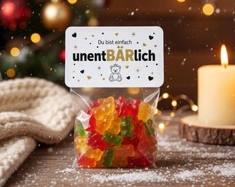 May include: A clear cellophane bag filled with colorful gummy bear candies. The bag has a white label with the text "Du bist einfach unentBÄRLich" and a teddy bear graphic. The background includes a lit candle and Christmas decorations.