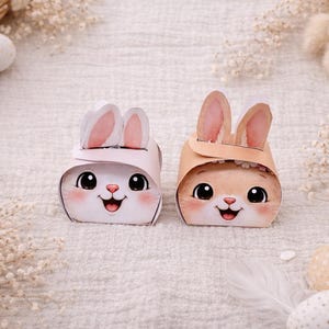 Puede incluir: Dos cajas de papel con forma de conejito de Pascua, una blanca y otra marrón claro, con orejas internas rosas y caras sonrientes. Las cajas están decoradas con caras de conejitos de dibujos animados. El fondo incluye huevos de Pascua, plumas y detalles florales.