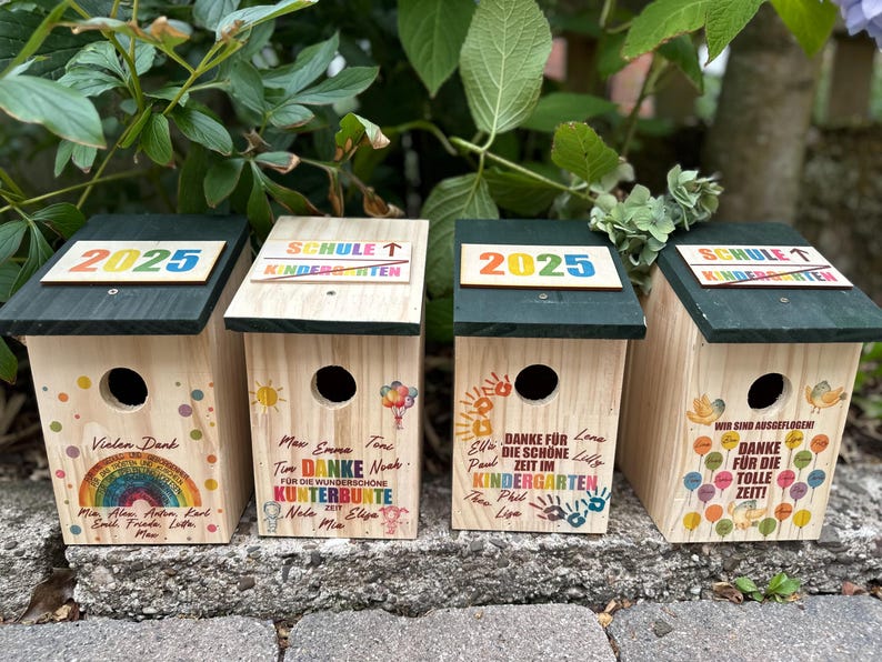 May include: Four wooden birdhouses with dark green roofs. Each birdhouse has a round entry hole and is decorated with colorful text and graphics. The text includes "2025", "SCHULE", and "KINDERGARTEN".