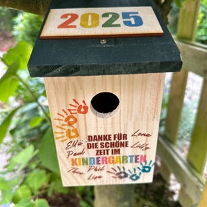 May include: A wooden birdhouse with a dark green roof and the year "2025" in colorful letters. The birdhouse has a round entry hole and text that reads "DANKE F&Uuml;R DIE SCH&Ouml;NE ZEIT IM KINDERGARTEN" with names and handprints.