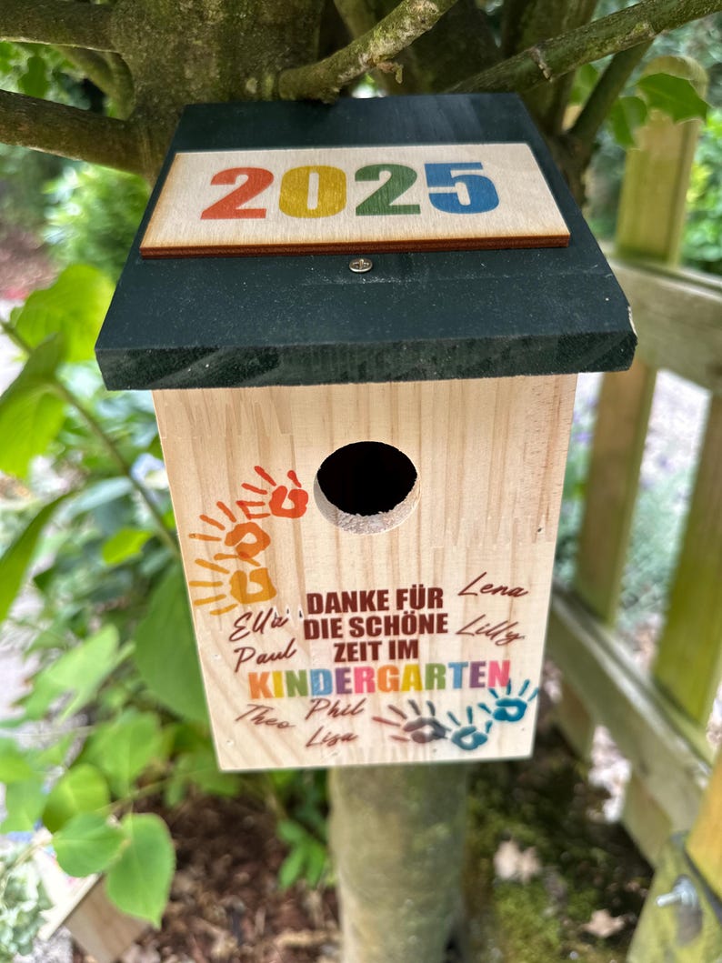 May include: A wooden birdhouse with a dark green roof and the year "2025" in colorful letters. The birdhouse has a round entry hole and text that reads "DANKE F&Uuml;R DIE SCH&Ouml;NE ZEIT IM KINDERGARTEN" with names and handprints.