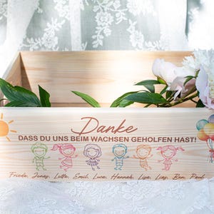 May include: Wooden box with a painted sun, children, and text in German. The box has a floral arrangement and balloons. The text says "Danke" and "Dass du uns beim Wachsen geholfen hast!" with names below.