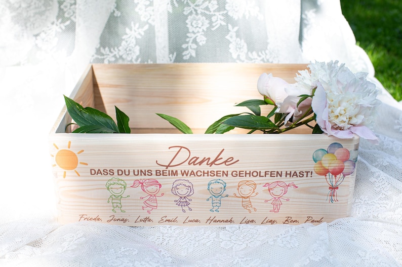 May include: Wooden box with a painted sun, children, and text in German. The box has a floral arrangement and balloons. The text says "Danke" and "Dass du uns beim Wachsen geholfen hast!" with names below.