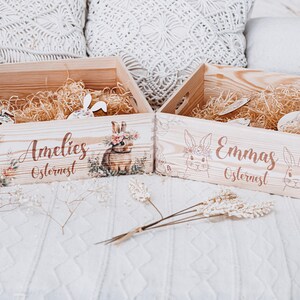 Puede incluir: Dos cestas de Pascua de madera llenas de paja y conejos decorativos de madera. Una cesta est&aacute; etiquetada "Amelies Osternest" con una ilustraci&oacute;n de conejo floral. La otra cesta est&aacute; etiquetada "Emmas Osternest".