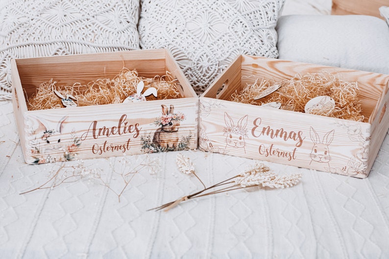 Puede incluir: Dos cestas de Pascua de madera llenas de paja y conejos decorativos de madera. Una cesta est&aacute; etiquetada "Amelies Osternest" con una ilustraci&oacute;n de conejo floral. La otra cesta est&aacute; etiquetada "Emmas Osternest".