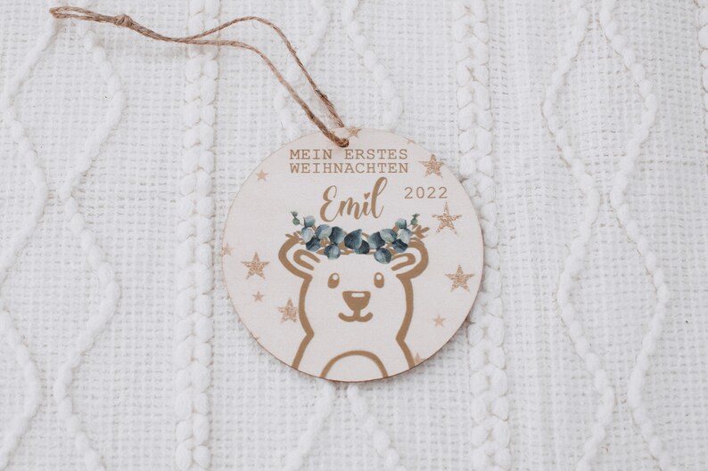 K&ouml;nnte beinhalten: Ein runder Holzanh&auml;nger mit einer wei&szlig;en B&auml;renillustration, goldenen Glitzer und dem Text "Mein erstes Weihnachten Emil 2022".