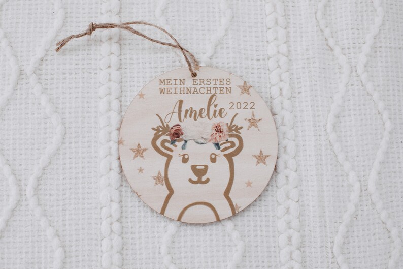 K&ouml;nnte beinhalten: Ein runder Holzanh&auml;nger mit goldfarbenem Glitzerhintergrund und einem wei&szlig;en B&auml;ren mit einer Blumenkrone. Der Anh&auml;nger tr&auml;gt die Aufschrift "Mein erstes Weihnachten" und "Amelie 2022".