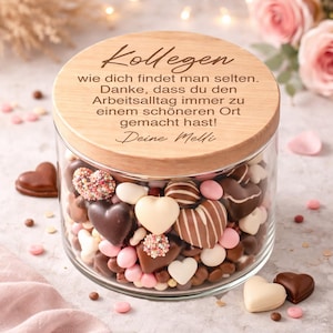 May include: A glass jar filled with heart-shaped chocolates and candies, topped with a wooden lid. The lid has German text. The chocolates are in various colors and designs, including white, brown, and pink.