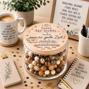 Peut inclure: Un bocal en verre transparent rempli de boules de chocolat marron et crème, surmonté d'un couvercle en bois avec du texte allemand, des cœurs et un motif de tasse à café. Le bocal est entouré d'un mug, de carnets et de stylos.