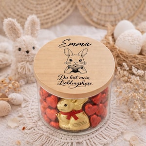 Puede incluir: Un tarro de cristal lleno de caramelos rojos en forma de corazón y un conejito de chocolate dorado. La tapa de madera está grabada con una ilustración de un conejo, el nombre "Emma" y la frase "Du bist mein Lieblingshase". Un conejito de peluche está en el fondo.