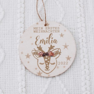 K&ouml;nnte beinhalten: Ein runder Holzanh&auml;nger mit einem goldenen Rentier-Motiv und dem Text "Mein erstes Weihnachten Emilia 2022".