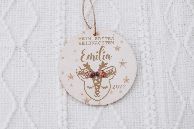K&ouml;nnte beinhalten: Ein runder Holzanh&auml;nger mit einem goldenen Rentier-Motiv und dem Text "Mein erstes Weihnachten Emilia 2022".