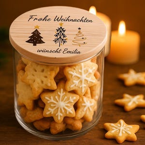 Op de afbeelding: Een glazen pot gevuld met stervormige koekjes, sommige versierd met witte glazuur. De houten deksel heeft de tekst "Frohe Weihnachten wünscht Emilia" en illustraties van kerstbomen. Brandende kaarsen op de achtergrond.