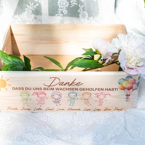 May include: A wooden box with the German word "Danke" and the phrase "DASS DU UNS BEIM WACHSEN GEHOLFEN HAST!" printed on the front. The box is decorated with cartoon figures, a sun, balloons, and names. White flowers and green leaves are inside.