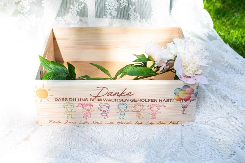 May include: A wooden box with the German word "Danke" and the phrase "DASS DU UNS BEIM WACHSEN GEHOLFEN HAST!" printed on the front. The box is decorated with cartoon figures, a sun, balloons, and names. White flowers and green leaves are inside.