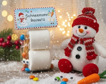 May include: A festive snowman kit with marshmallows, pretzel sticks, and candy eyes in a clear bag. A plush snowman with a red hat and scarf sits nearby, along with a carrot nose and red ball feet. The bag has the text "Schneemann Bausatz".