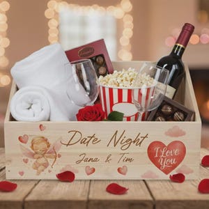 Könnte beinhalten: Eine Holzkiste mit einer Weinflasche, Pralinen, Popcorn, zwei Weingläsern und einem gerollten weißen Handtuch. Die Kiste ist mit einem Herz, einem Engel und den Worten "Date Night" und "I Love You" verziert.