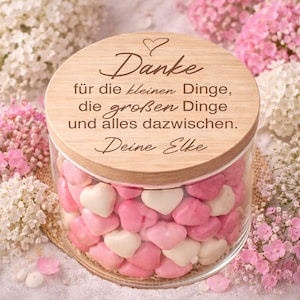 Op de afbeelding: Een glazen pot gevuld met roze en witte hartvormige snoepjes. De pot heeft een houten deksel met het woord "Danke" en andere tekst gegraveerd. De pot is omgeven door roze en witte bloemen.