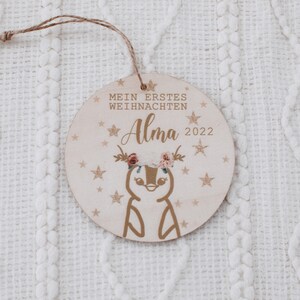 K&ouml;nnte beinhalten: Ein runder Holzanh&auml;nger mit goldfarbenem Glitzerhintergrund und einer wei&szlig;en Pinguin-Illustration. Der Anh&auml;nger tr&auml;gt den Text "Mein erstes Weihnachten Alma 2022".