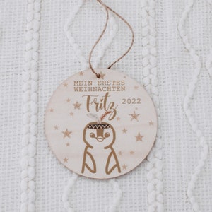 K&ouml;nnte beinhalten: Ein Holz-Weihnachtsornament mit einem Pinguin, der ein Kopfschmuck tr&auml;gt und den Text "Mein erstes Weihnachten Fritz 2022" umgeben von Sternen.