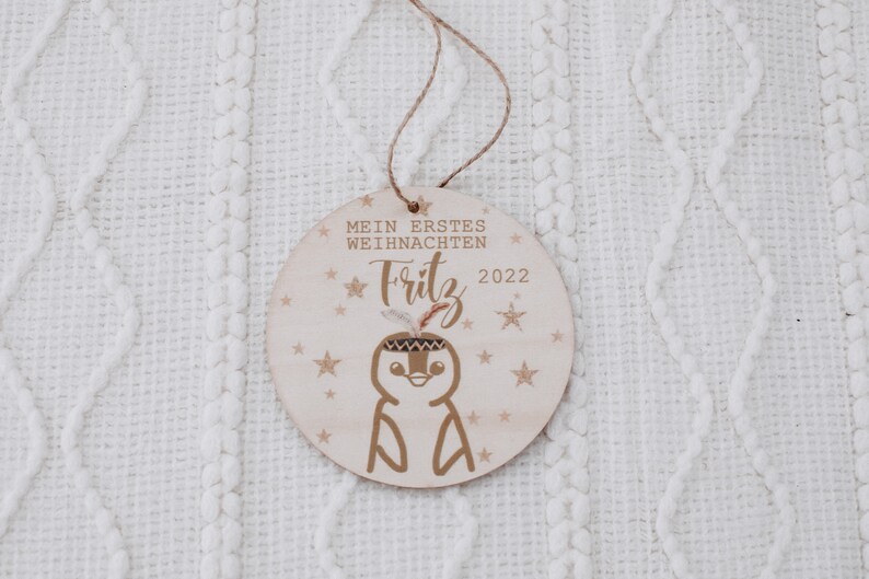 K&ouml;nnte beinhalten: Ein Holz-Weihnachtsornament mit einem Pinguin, der ein Kopfschmuck tr&auml;gt und den Text "Mein erstes Weihnachten Fritz 2022" umgeben von Sternen.