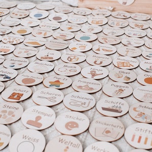 May include: A set of 60 wooden circles with various illustrations and German words printed on them. The circles are beige with brown accents and feature illustrations of objects, activities, and concepts.