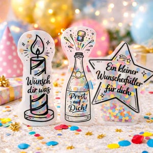 May include: Three decorative party favors. One is shaped like a candle with the text "Wünsch dir was". Another is a champagne bottle with the text "Prost auf Dich!". The third is a star with the text "Ein kleiner Wunschfüller für dich". All are filled with confetti.