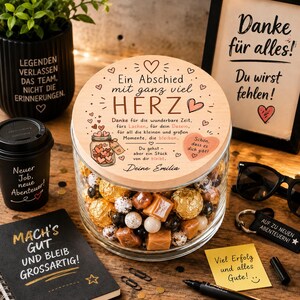 Könnte beinhalten: Ein klares Glasgefäß gefüllt mit gemischten Süßigkeiten, mit einem runden Holzdeckel, der mit deutschem Text wie "Ein Abschied mit ganz viel HERZ" und einem Herzmotiv graviert ist, umgeben von verschiedenen Büro-Geschenkartikeln.