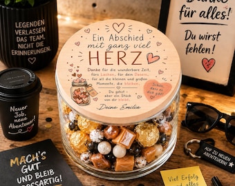 Puede incluir: Un frasco de vidrio transparente lleno de dulces variados, con una tapa de madera redonda grabada con texto en alemán que incluye "Ein Abschied mit ganz viel HERZ" y un corazón, rodeado de varios artículos de regalo de oficina.