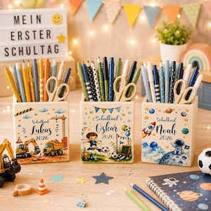 Könnte beinhalten: Drei quadratische Stiftehalter aus Holz, jeweils personalisiert mit einem Namen und dem Jahr 2026. Der erste zeigt Baufahrzeuge, der zweite einen Fußballspieler und der dritte ein Weltraumthema mit Rakete und Astronaut, alle gefüllt mit Stiften.
