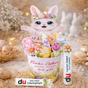 Può includere: Una composizione decorativa a tema pasquale con un coniglietto di carta con una corona floreale, uova di carta, fiori e coniglietti in un secchio decorativo. Il secchio ha testo in tedesco. È presente anche un bastoncino bianco con testo in tedesco.