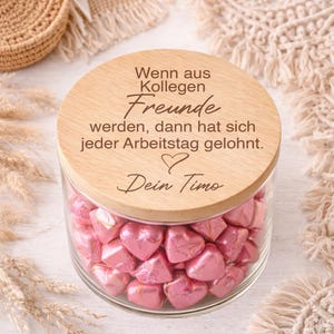 Könnte beinhalten: Ein klares Glas mit rosa herzförmigen Süßigkeiten, bedeckt mit einem Holzdeckel. Der Deckel hat deutschen Text. Das Glas ist von dekorativen Elementen umgeben.