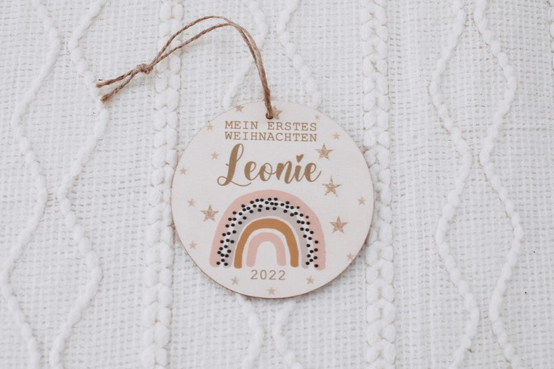 K&ouml;nnte beinhalten: Ein runder Holzanh&auml;nger mit einem Regenbogen, Sternen und dem Text "Mein erstes Weihnachten Leonie 2022".