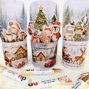 Może przedstawiać: Trzy świąteczne dekoracje bożonarodzeniowe z zimowymi scenami i postaciami. Każda ma inny wzór, w tym Świętego Mikołaja, elfy i piernikowych ludzików. Dekoracjom towarzyszą batony czekoladowe.