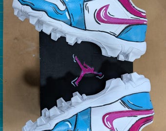 May include: Pair of white and blue athletic cleats with a pink swoosh design. The cleats have a cartoon-like aesthetic with black outlines. The shoes are placed on top of a black box with a pink Jumpman logo.