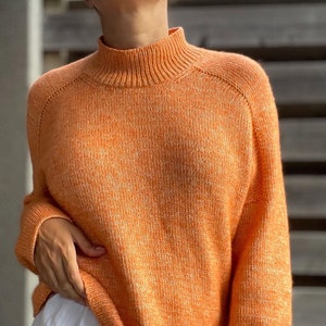 May include: A soft, orange knit sweater with a mock neck and long sleeves. The sweater has a slightly cropped length and a relaxed fit.