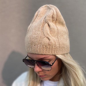 Può includere: Un berretto di lana beige con un motivo a treccia e un bordo ripiegato.