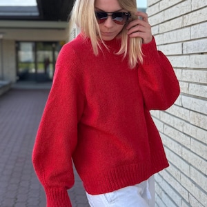 Può includere: Un maglione rosso brillante a maglia con collo alto e maniche lunghe. Il maglione ha orlo e polsini a coste. La persona indossa pantaloni bianchi e occhiali da sole.