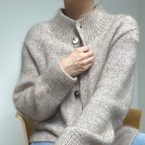 Peut inclure: Un cardigan beige à boutons avec un col montant. Le pull est fait d'un tissu tricoté texturé et comporte des poignets côtelés et une fermeture à boutons. Les manches sont longues et le pull semble chaud et confortable.