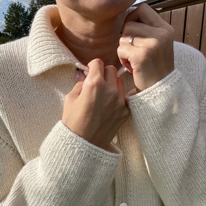 May include: Cream-colored, knitted cardigan with a collared neckline and button-down front. The sleeves have ribbed cuffs. The cardigan is made of a thick, textured yarn. The buttons are white and round.