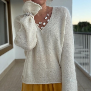 Puede incluir: Suéter crema con cuello en V y mangas largas con puños con volantes. Se ve un collar con colgantes blancos redondos grandes y una pulsera con detalles blancos y plateados. También se ve una falda amarilla.